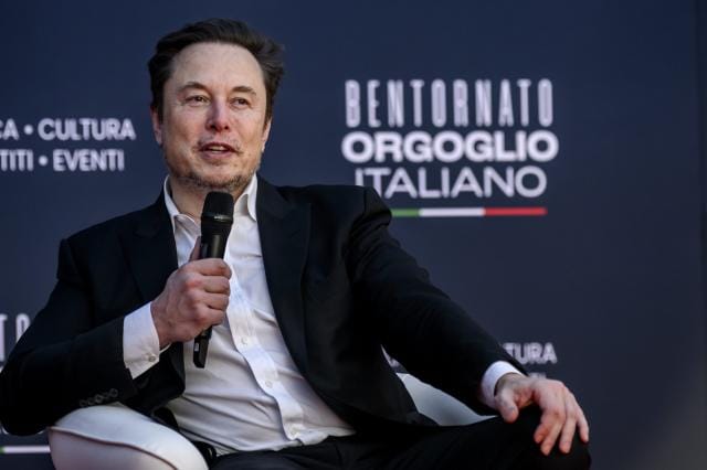 Elon Musk sitting on stage holding a microphone.