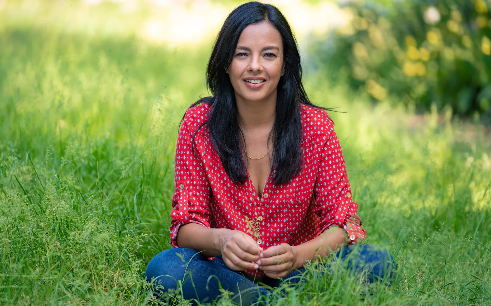 Liz Bonnin sitting on the grass wearing a red shirt and jeans.
