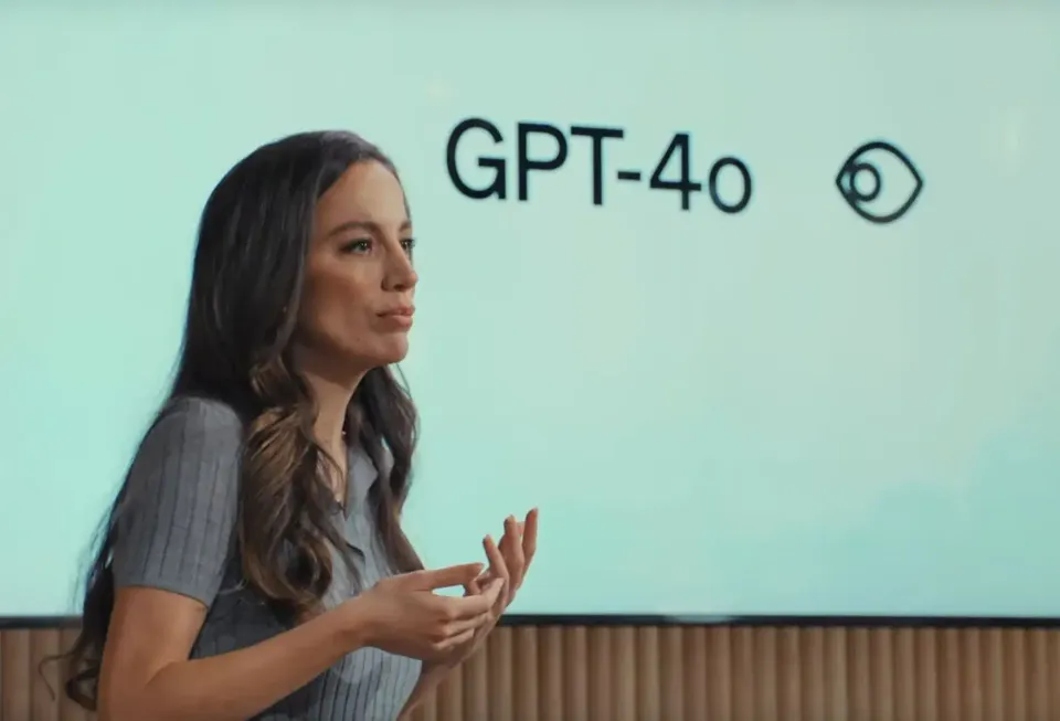 Close shot of woman at front of room giving presentation. GPT-4o is on a projection board behind her.